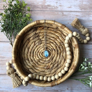 May include: A silver-toned pendant necklace featuring an oval-shaped charm with a light blue background and dark text. The pendant hangs from a delicate silver chain, centred within a round, woven tan wicker basket with a natural texture.