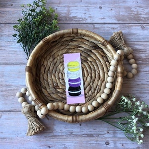 Puede incluir: Un marcapáginas rectangular con una pila vertical de cuatro macarons en amarillo, blanco, morado y negro sobre un fondo morado claro. El marcapáginas está centrado dentro de una cesta redonda de hierba marina tejida con una guirnalda de cuentas.