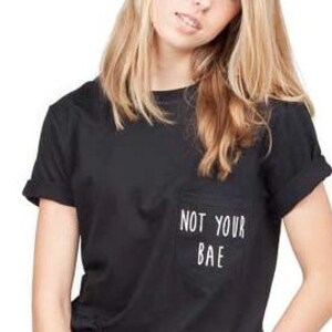Puede incluir: Una camiseta negra con un bolsillo blanco que dice "Not Your Bae".