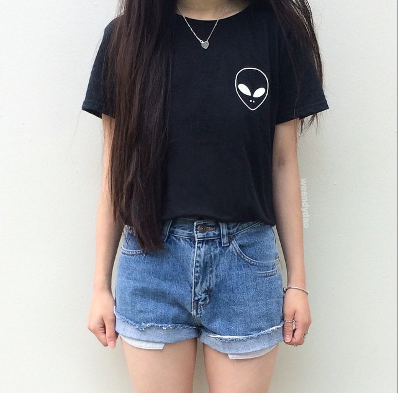 May include: A black t-shirt with a white alien face graphic on the front. The shirt is paired with blue denim shorts.