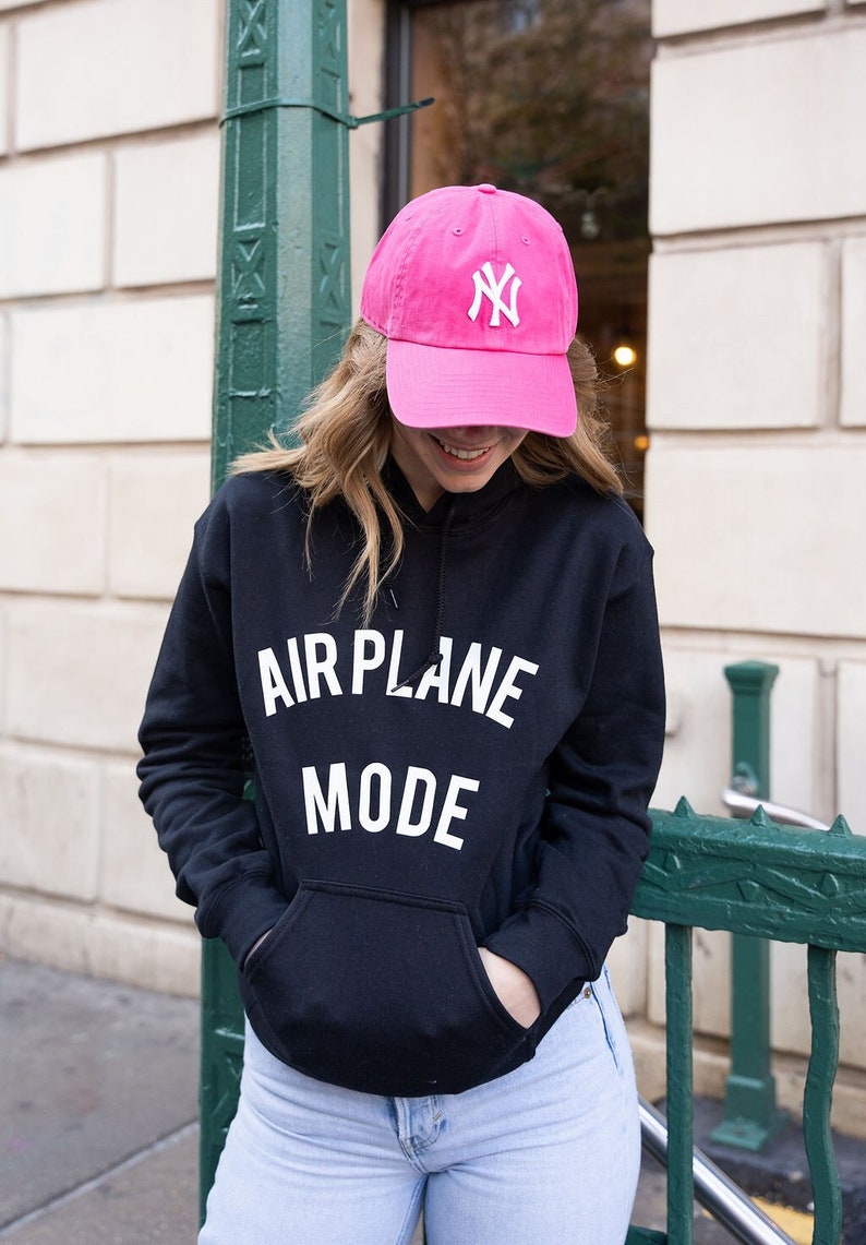 Może przedstawiać: Czarna bluza z kapturem z napisem "AIRPLANE MODE" w białych literach. Osoba nosi r&oacute;żową czapkę bejsbolową z białym logo "NY".