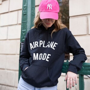 Może przedstawiać: Czarny sweterek z kapturem z napisem "AIRPLANE MODE" w białym kolorze. Osoba nosi r&oacute;żową czapkę z daszkiem z białym logo "NY".