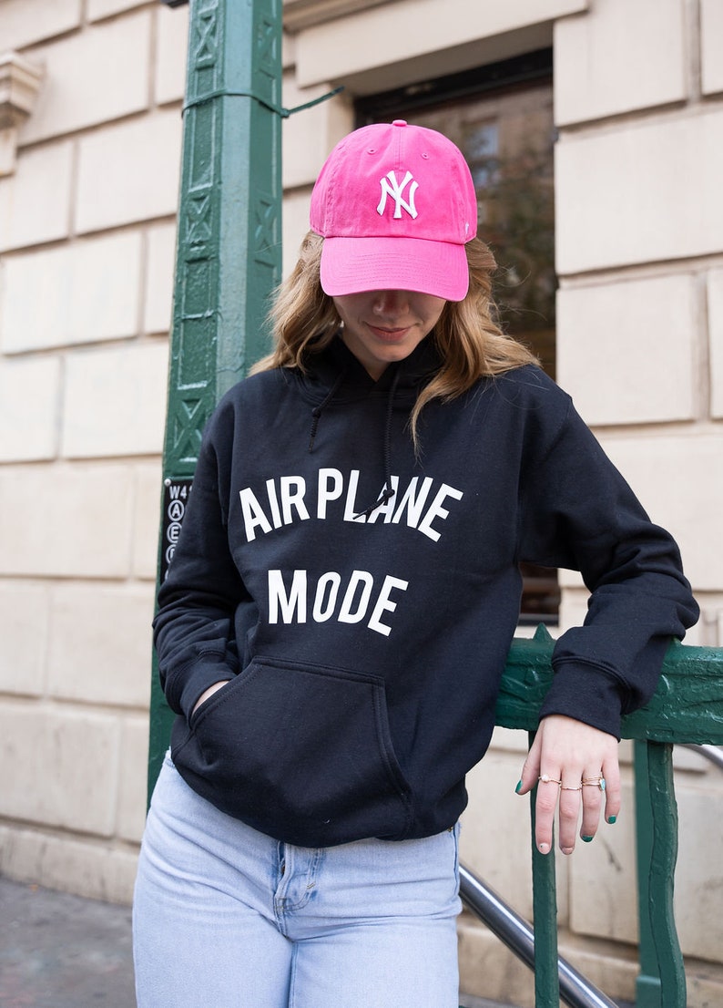 Może przedstawiać: Czarny sweterek z kapturem z napisem "AIRPLANE MODE" w białym kolorze. Osoba nosi r&oacute;żową czapkę z daszkiem z białym logo "NY".