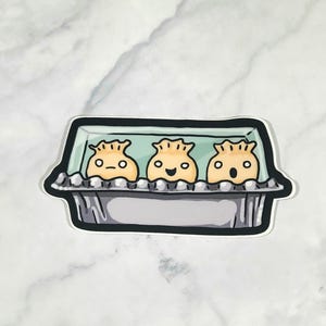 May include: A sticker featuring three cartoon dumplings in a foil container. The dumplings have smiling faces and are colored in shades of yellow and orange. The container is silver with a black outline, set against a white and gray marble background.