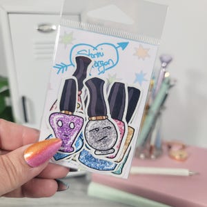 Puede incluir: Un paquete transparente con pegatinas de esmalte de u&ntilde;as. Las pegatinas presentan dibujos de botes de esmalte de u&ntilde;as en varios colores, incluyendo morado, plateado y rosa, con caras expresivas. El paquete tiene el texto "Stormi Morgan" en un dise&ntilde;o de coraz&oacute;n.