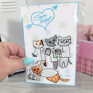 May include: A clear plastic package containing a set of stickers featuring cartoon cats dressed as scientists. The stickers have a white background with a blue heart and arrow design with the text "Stormi Morgan". The cats are in lab coats and holding lab equipment.