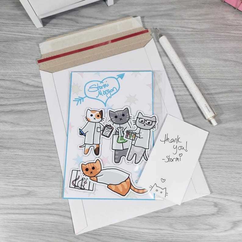 May include: A white envelope with a clear plastic sleeve containing a card with cartoon cats in lab coats. The card has the text "Storm Morgan" and "Thank you! -Storm!" written on it. A white pen is also included.