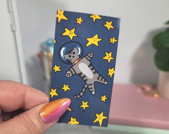 May include: A rectangular sticker featuring a gray tabby cat in a space helmet floating among yellow stars on a navy blue background. The cat is wearing a red collar. The sticker is held by a hand with a colorful manicure.