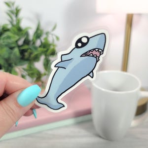 May include: A light blue cartoon shark sticker with black outlines and large, round eyes. The shark has a white mouth with pink gums and teeth. The sticker is held in front of a white mug and a small green plant.