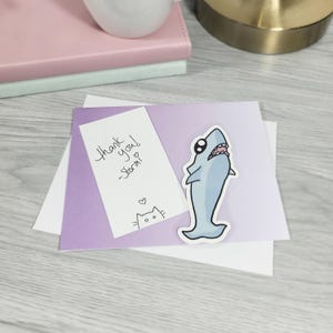 May include: A light blue shark sticker with a cartoon design, placed on a purple and white card. A handwritten note says "Thank you! -Stormi" with a cat drawing. The card is on a wooden surface.