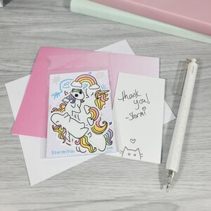 May include: A collection of stationery items including a pink envelope, a white card with the text "Thank you! -Stormi" and a sticker sheet with unicorn illustrations. A white pen is also visible.