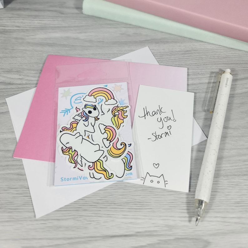 May include: A collection of stationery items including a pink envelope, a white card with the text "Thank you! -Stormi" and a sticker sheet with unicorn illustrations. A white pen is also visible.