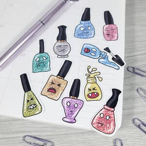 Puede incluir: Una colecci&oacute;n de pegatinas de esmalte de u&ntilde;as de colores con varios acabados brillantes y caras de dibujos animados. Las pegatinas son en tonos de azul, rosa, verde, dorado y morado. Tambi&eacute;n se ven un bol&iacute;grafo y clips.