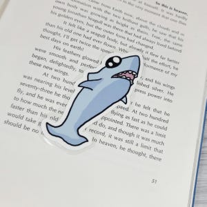 May include: A light blue shark sticker with a cartoon design. The sticker has a white outline and a smiling mouth. The sticker is placed on top of an open book.