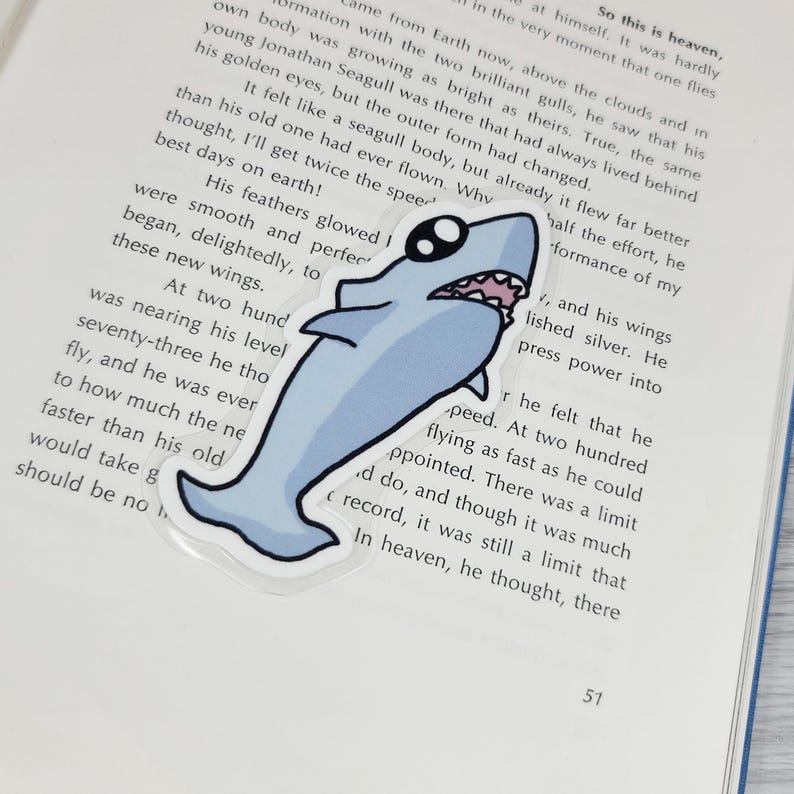 May include: A light blue shark sticker with a cartoon design. The sticker has a white outline and a smiling mouth. The sticker is placed on top of an open book.