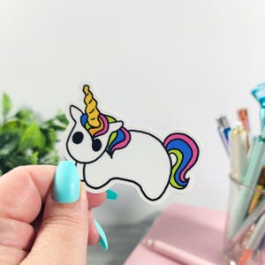May include: A white unicorn sticker with a black outline, featuring a yellow horn, a rainbow mane, and a colorful tail. The sticker is held by a hand with blue nail polish.