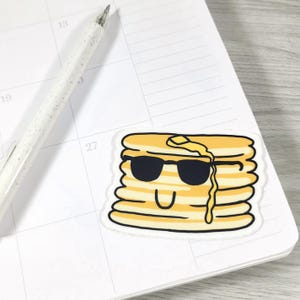 May include: A sticker featuring a stack of pancakes with black sunglasses and a smiling face. The pancakes are yellow with a white outline, and a pat of butter is melting on top. The sticker is on a white calendar page.