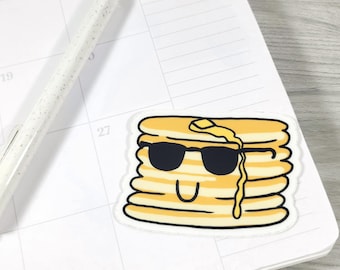 May include: A sticker featuring a stack of pancakes with black sunglasses and a smiling face. The pancakes are yellow with a white outline, and a pat of butter is melting on top. The sticker is on a white calendar page.