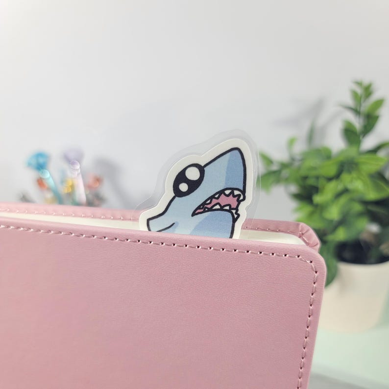 May include: A shark-shaped bookmark peeks out from a pink leather-bound notebook. The cartoon shark is light blue with a white belly, black eyes, and a pink mouth. The bookmark is made of clear plastic.