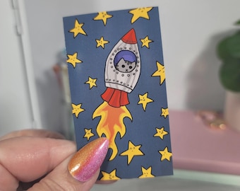 May include: A rectangular sticker featuring a cartoon rocket ship with a cat in the cockpit, surrounded by yellow stars on a dark blue background. The rocket has red and silver details, with orange flames. The cat has gray fur.