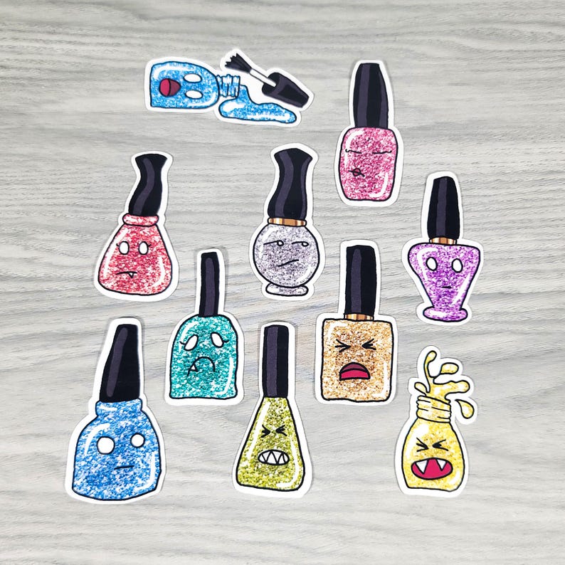 Puede incluir: Una colecci&oacute;n de pegatinas de dibujos animados de esmalte de u&ntilde;as en varios colores, incluyendo rosa, azul y dorado, cada una con una expresi&oacute;n facial &uacute;nica. Una pegatina muestra esmalte derramado. Las pegatinas tienen tapas negras.