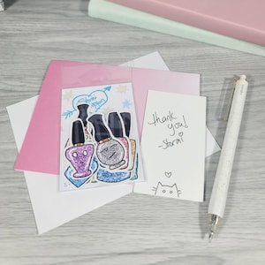 Puede incluir: Una colecci&oacute;n de pegatinas de esmalte de u&ntilde;as con caras de dibujos animados, un sobre rosa, una tarjeta blanca con "Thank you! -Stormi" escrito y un bol&iacute;grafo blanco. Las pegatinas est&aacute;n en una bolsa de pl&aacute;stico transparente.
