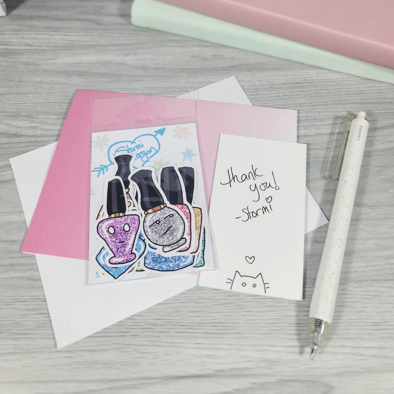 Puede incluir: Una colecci&oacute;n de pegatinas de esmalte de u&ntilde;as con caras de dibujos animados, un sobre rosa, una tarjeta blanca con "Thank you! -Stormi" escrito y un bol&iacute;grafo blanco. Las pegatinas est&aacute;n en una bolsa de pl&aacute;stico transparente.