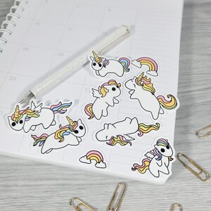 May include: A collection of white unicorn stickers with colorful manes, horns, and wings, along with rainbow stickers. The stickers are scattered on a calendar page next to a pen and paperclips. The unicorns have yellow, pink, and blue accents.