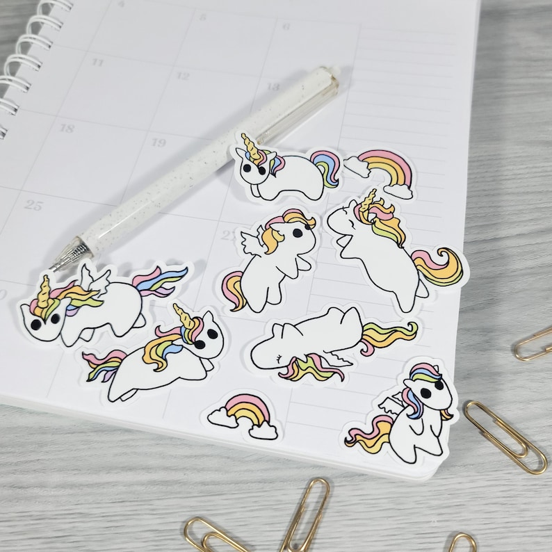 May include: A collection of white unicorn stickers with colorful manes, horns, and wings, along with rainbow stickers. The stickers are scattered on a calendar page next to a pen and paperclips. The unicorns have yellow, pink, and blue accents.