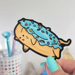 May include: A sticker featuring a cartoon cat in the shape of a donut. The donut has blue frosting with colorful sprinkles. The cat is light brown with black details. The sticker is on a white background.