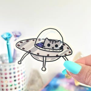 May include: A sticker featuring a gray tabby cat piloting a silver spaceship. The spaceship has red lights and a clear dome. The cat is looking out from the spaceship. The sticker is held by a hand with blue fingernails.