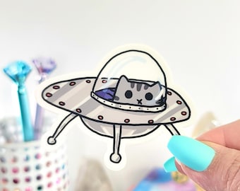 May include: A sticker featuring a gray tabby cat piloting a silver spaceship. The spaceship has red lights and a clear dome. The cat is looking out from the spaceship. The sticker is held by a hand with blue fingernails.