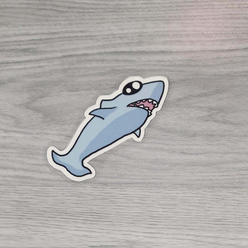 May include: A light blue shark sticker with black sunglasses and a white border. The shark has a wide open mouth with pink gums and white teeth. The sticker is on a light gray wooden surface.