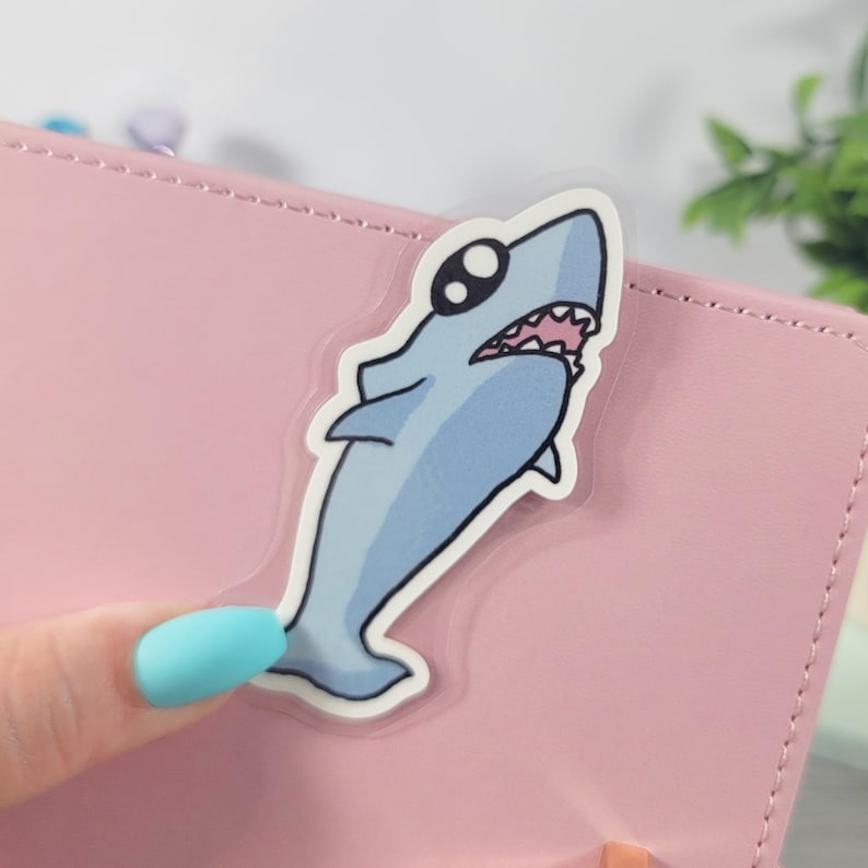 May include: A light blue cartoon shark sticker with a white outline is placed on a pink surface. The shark has a wide open mouth with pink teeth and black eyes. The sticker is held by a person with a light blue fingernail.
