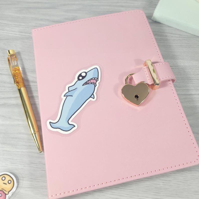 May include: Pink faux leather journal with a heart-shaped lock and a gold pen. A cartoon shark sticker is on the cover. The journal has a gold clasp and a stitched border. The pen has a gold barrel with glitter.