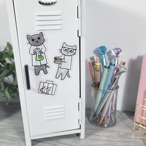 May include: White locker with cat-themed stickers. One sticker shows a cat in a lab coat holding a flask. Another sticker shows a cat with glasses holding test tubes. A glass jar holds pens with diamond-shaped tops.