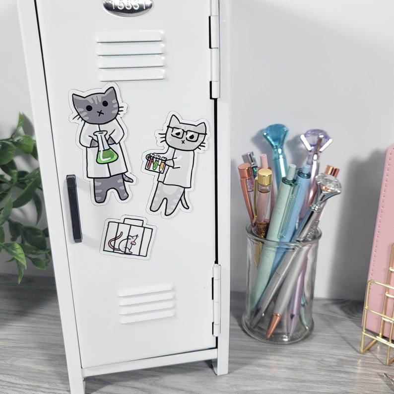 May include: White locker with cat-themed stickers. One sticker shows a cat in a lab coat holding a flask. Another sticker shows a cat with glasses holding test tubes. A glass jar holds pens with diamond-shaped tops.
