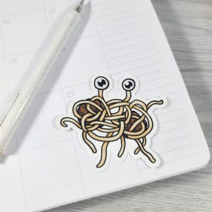 May include: A sticker featuring a cartoon depiction of the Flying Spaghetti Monster, with two googly eyes and spaghetti-like appendages. The sticker is on a white calendar page, with a pen in the background.