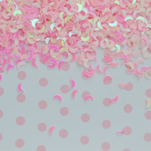 May include: Pink iridescent glitter in the shape of crescent moons scattered on a white background with pink polka dots.