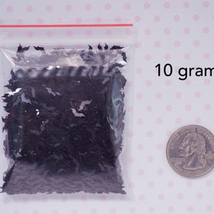 May include: A clear plastic bag filled with black glitter shaped like bats. The bag is sitting on a white surface with pink polka dots. A US quarter is shown for scale. 10 grams.