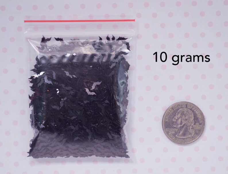 May include: A clear plastic bag filled with black glitter shaped like bats. The bag is sitting on a white surface with pink polka dots. A US quarter is shown for scale. 10 grams.