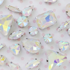 May include: A collection of iridescent, faceted glass rhinestones in various shapes including round, pear, heart, and rectangular cuts. Each clear crystal features a metallic silver prong setting along the edges, reflecting rainbow light.