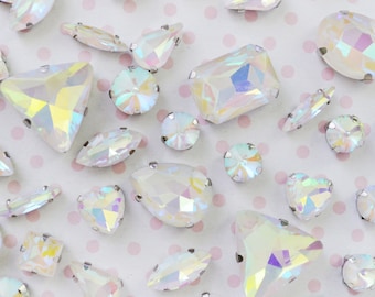 May include: Assortment of iridescent rhinestones in various shapes, including round, teardrop, and square. Each rhinestone has a silver-colored metal setting. The rhinestones are scattered on a light pink background with small pink polka dots.