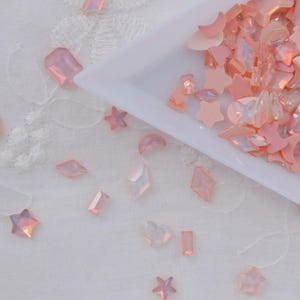 May include: A collection of pink and iridescent craft embellishments in various shapes, including stars, moons, diamonds, and rectangles. The embellishments are scattered on a white surface and in a white tray.