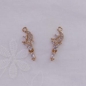 May include: Two gold-colored crescent moon charms with clear crystal accents. Each charm features a crescent moon shape with a cluster of crystals and a dangling rectangular crystal. The charms are designed for jewelry making.