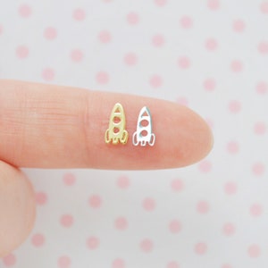 May include: A pair of small rocket ship earrings, one gold and one silver, shown on a finger.