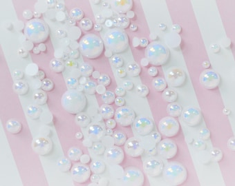 May include: A collection of round, white, iridescent cabochons of varying sizes scattered across a surface with light pink and white vertical stripes. Each cabochon features a smooth, domed top that reflects light with a subtle rainbow sheen.