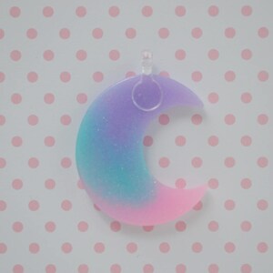 May include: A crescent moon pendant featuring a gradient of purple, teal, and pink colors with a subtle glitter finish. The moon shape is made of a smooth material and includes a clear plastic loop at the top for attachment to a chain or cord.
