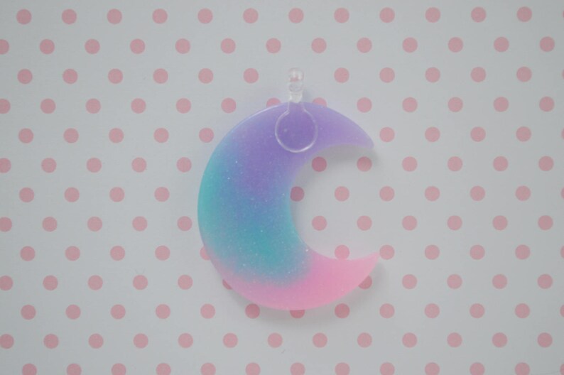 May include: A crescent moon pendant featuring a gradient of purple, teal, and pink colors with a subtle glitter finish. The moon shape is made of a smooth material and includes a clear plastic loop at the top for attachment to a chain or cord.
