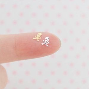 May include: Two tiny skull and crossbones charms, one gold and one silver, are shown on a fingertip.